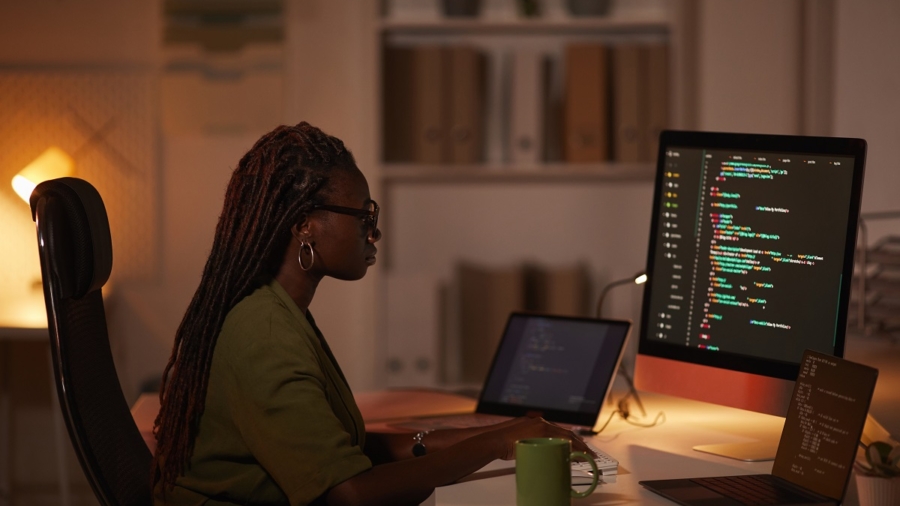 Side view portrait of contemporary African-American woman writing code and looking at computer screen while working in dim office, copy space
